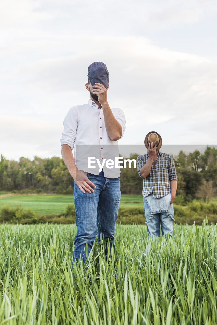 Farm worker covering face with hats while standing on farm against sky during sunset