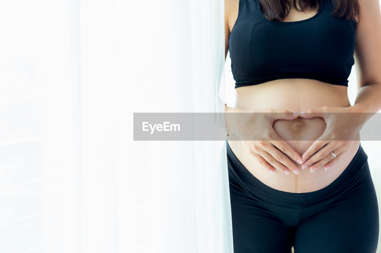 MIDSECTION OF YOUNG WOMAN STANDING AGAINST WALL AT HOME