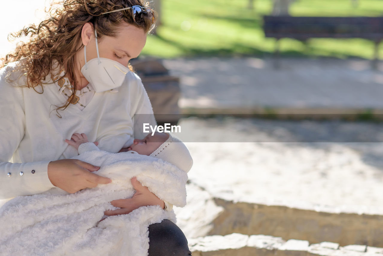 Mother wearing mask breastfeeding baby whiles sitting outdoors