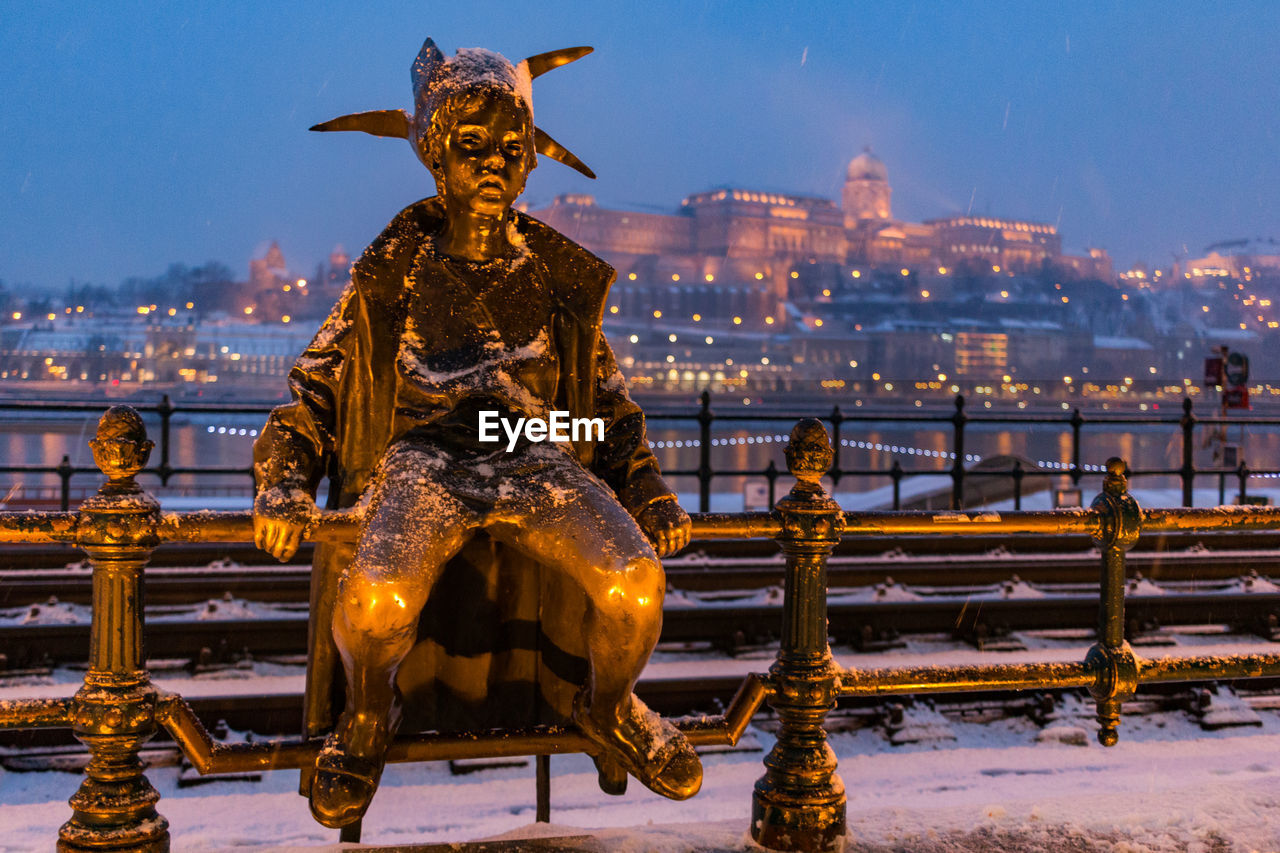 Statue by snow covered sidewalk near river in city at dusk