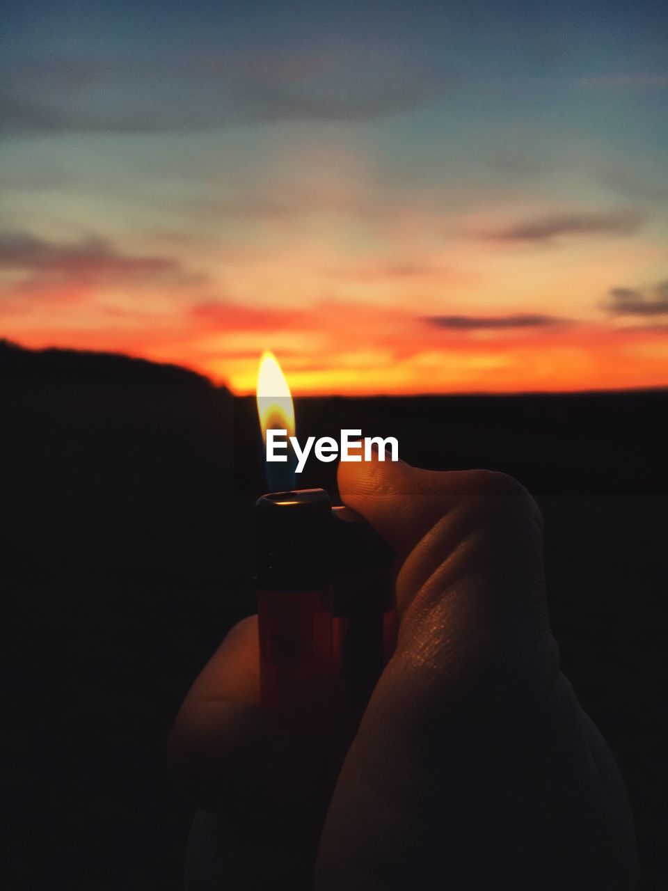 Cropped hand holding cigarette lighter against sky during sunset