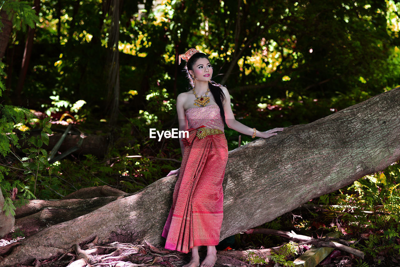 Young woman in traditional clothing looking away while standing by tree trunk at forest