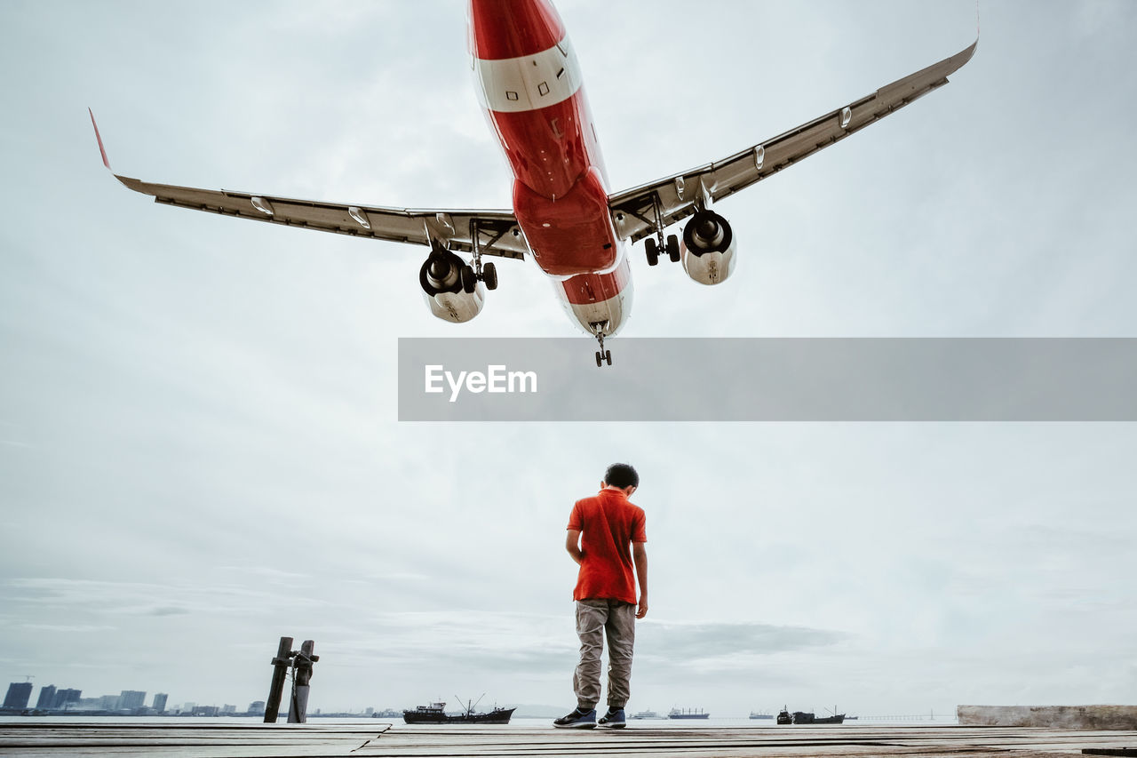 Rear view of boy standing on pier against airplane flying in sky
