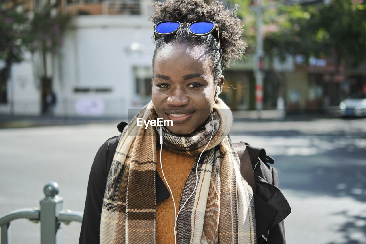 Young happy trendy african american female with rucksack and sunglasses strolling on urban roadway while looking at camera in sunlight