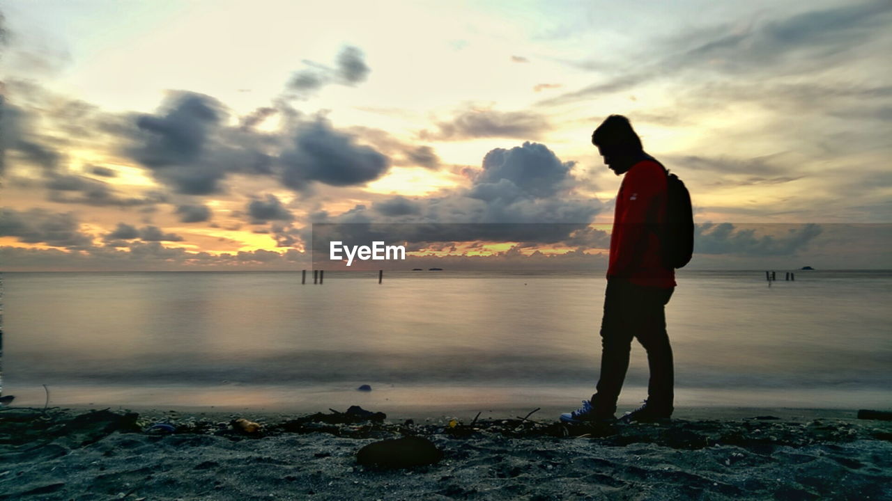 Side view of man standing at beach against sky during sunset