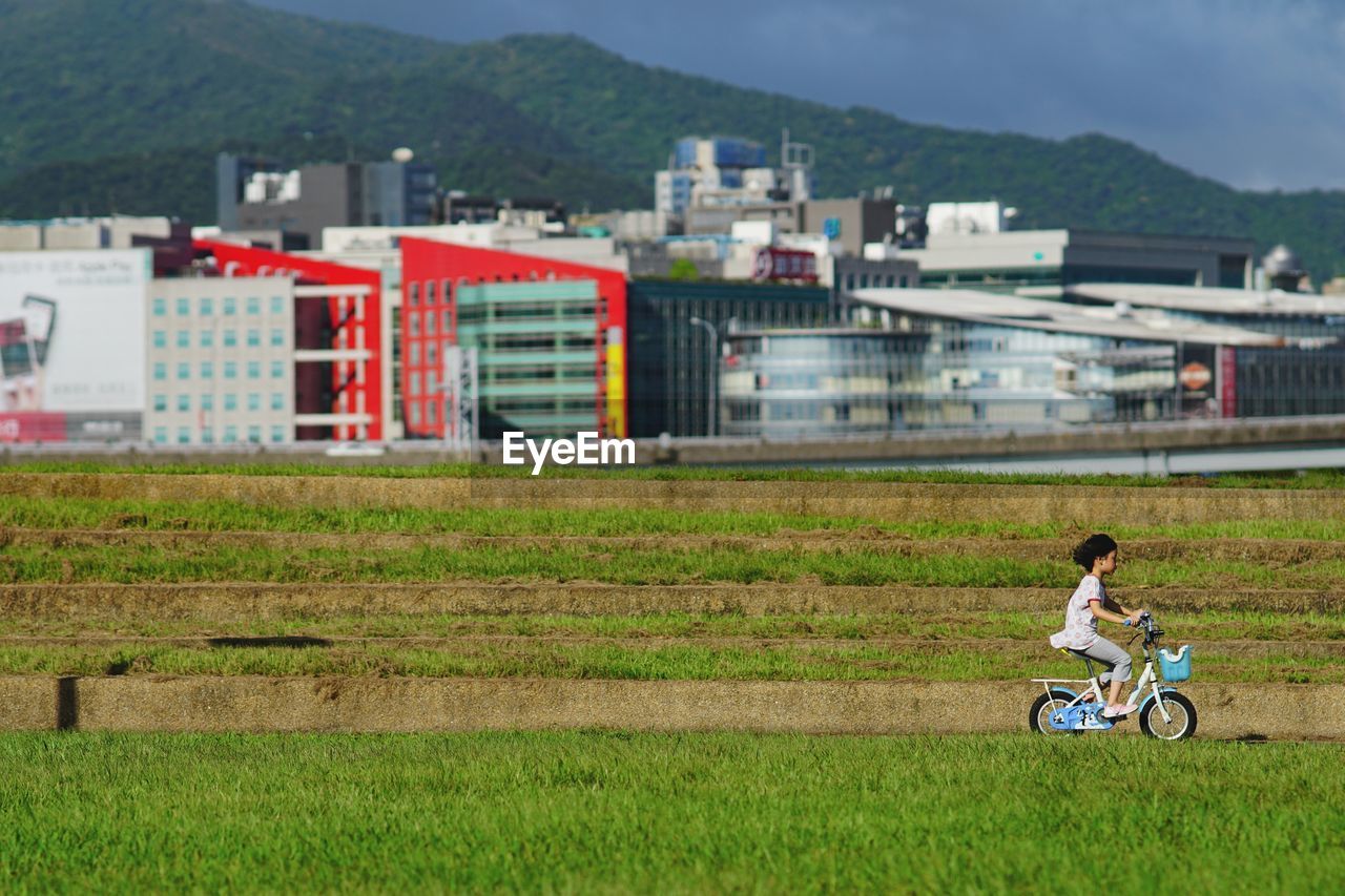 Side view of girl riding bicycle on grassy field against buildings