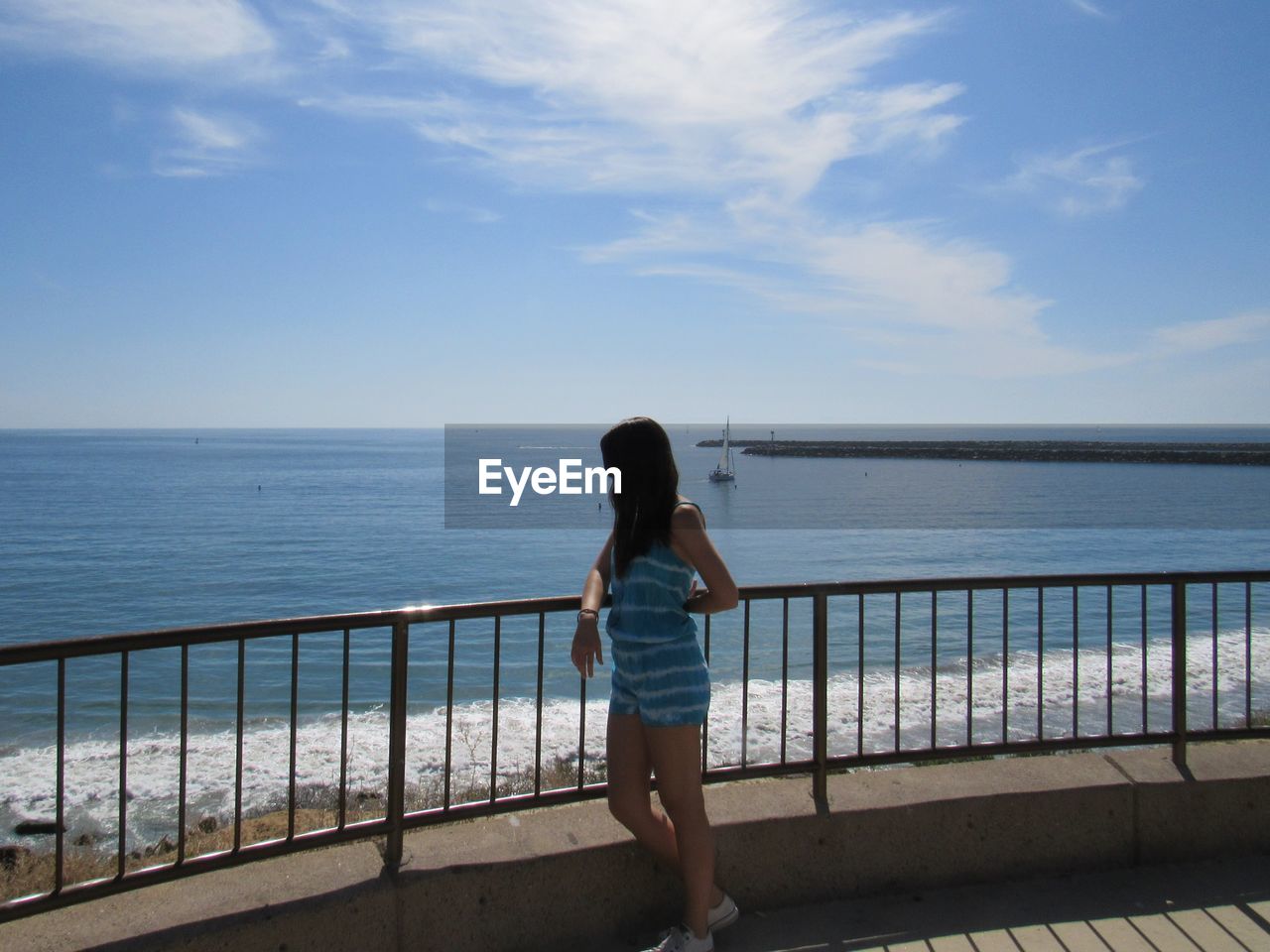 REAR VIEW OF WOMAN STANDING ON RAILING BY SEA AGAINST SKY