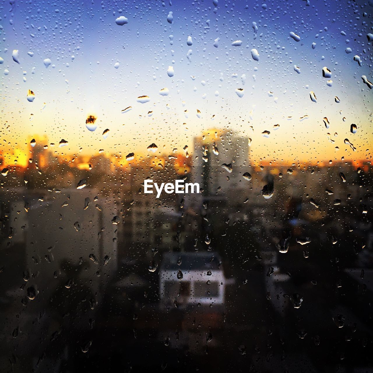 Buildings against sky seen through wet glass window at dusk