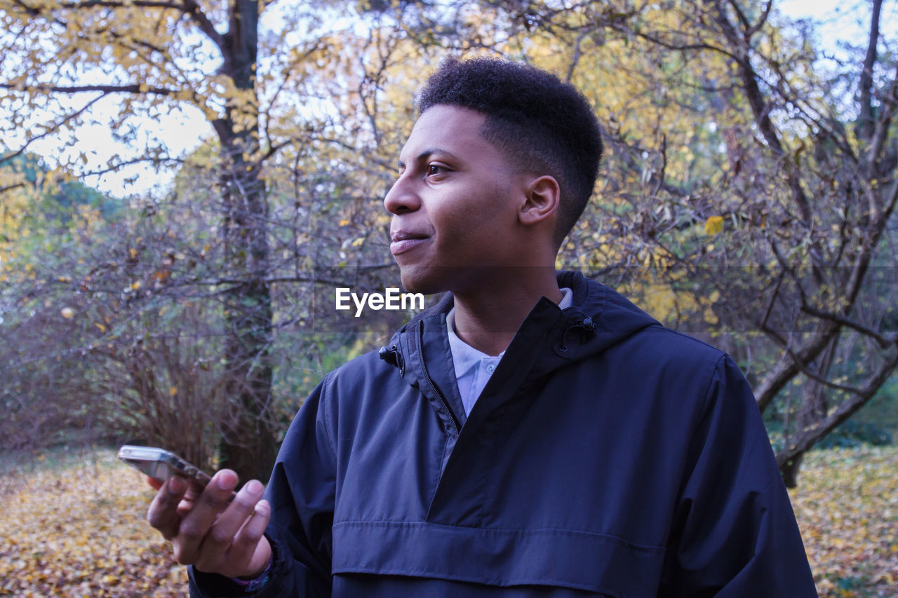 Young man looking away while holding mobile phone against tree