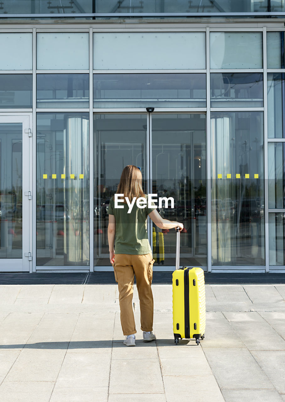 Young woman traveler carrying yellow suitcase next to the entrance to the airport outside tourism 