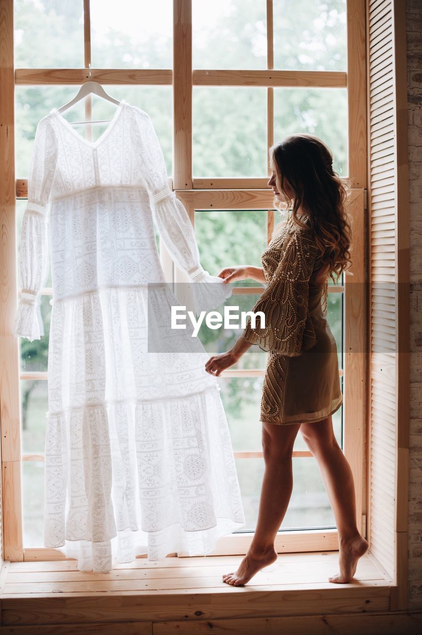 Side view of woman looking at white dress hanging on window at home