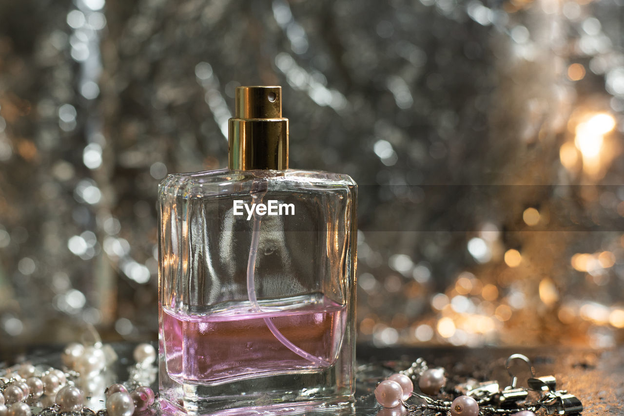 A square bottle of pink perfume on a wet floor, with orange and silver bokeh behind it