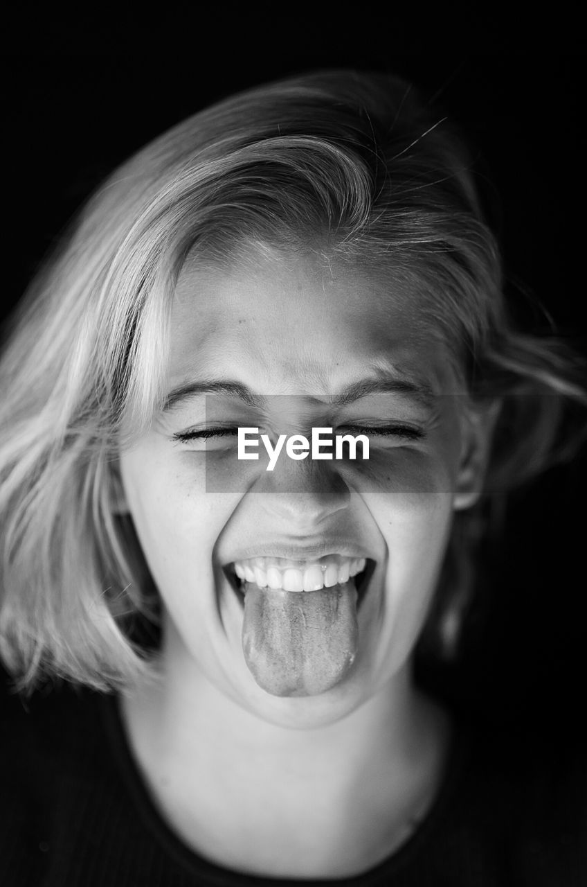 Close-up of young woman with eyes closed sticking out tongue against black background