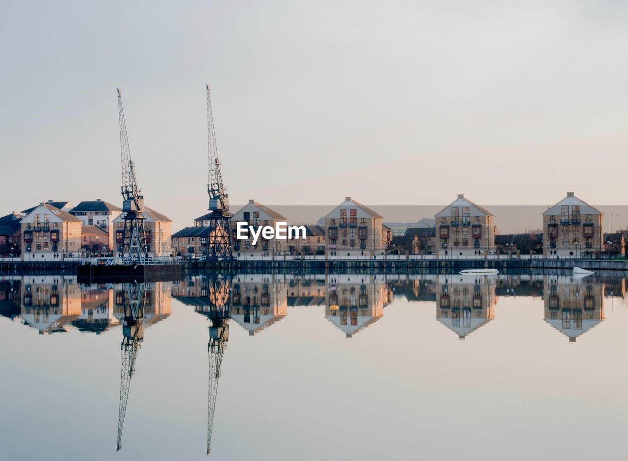 Reflection of buildings and cranes on lake against clear sky