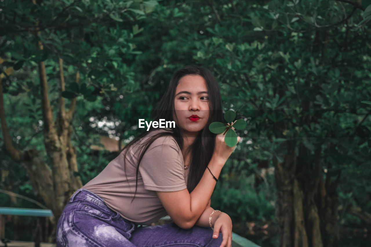 PORTRAIT OF BEAUTIFUL WOMAN HOLDING PLANT AGAINST TREES