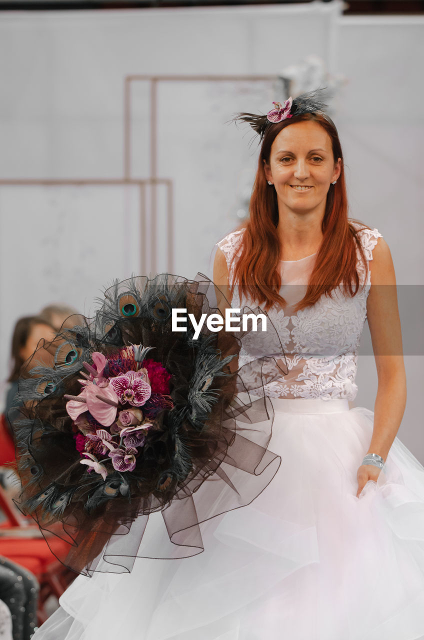 PORTRAIT OF BEAUTIFUL YOUNG WOMAN WITH FLOWER BOUQUET AGAINST WALL