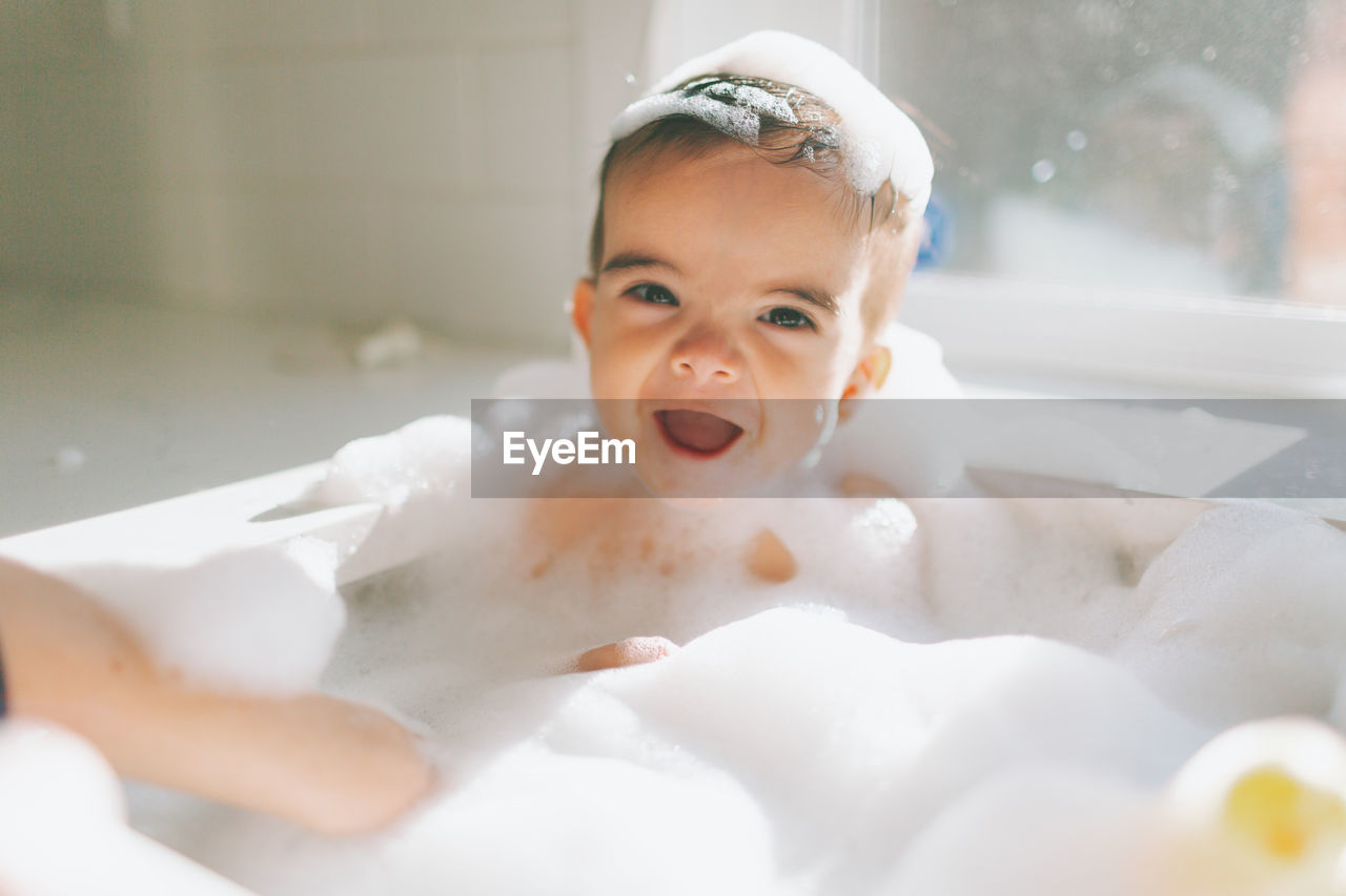 Cute happy baby boy taking bubble bath in kitchen sink at home