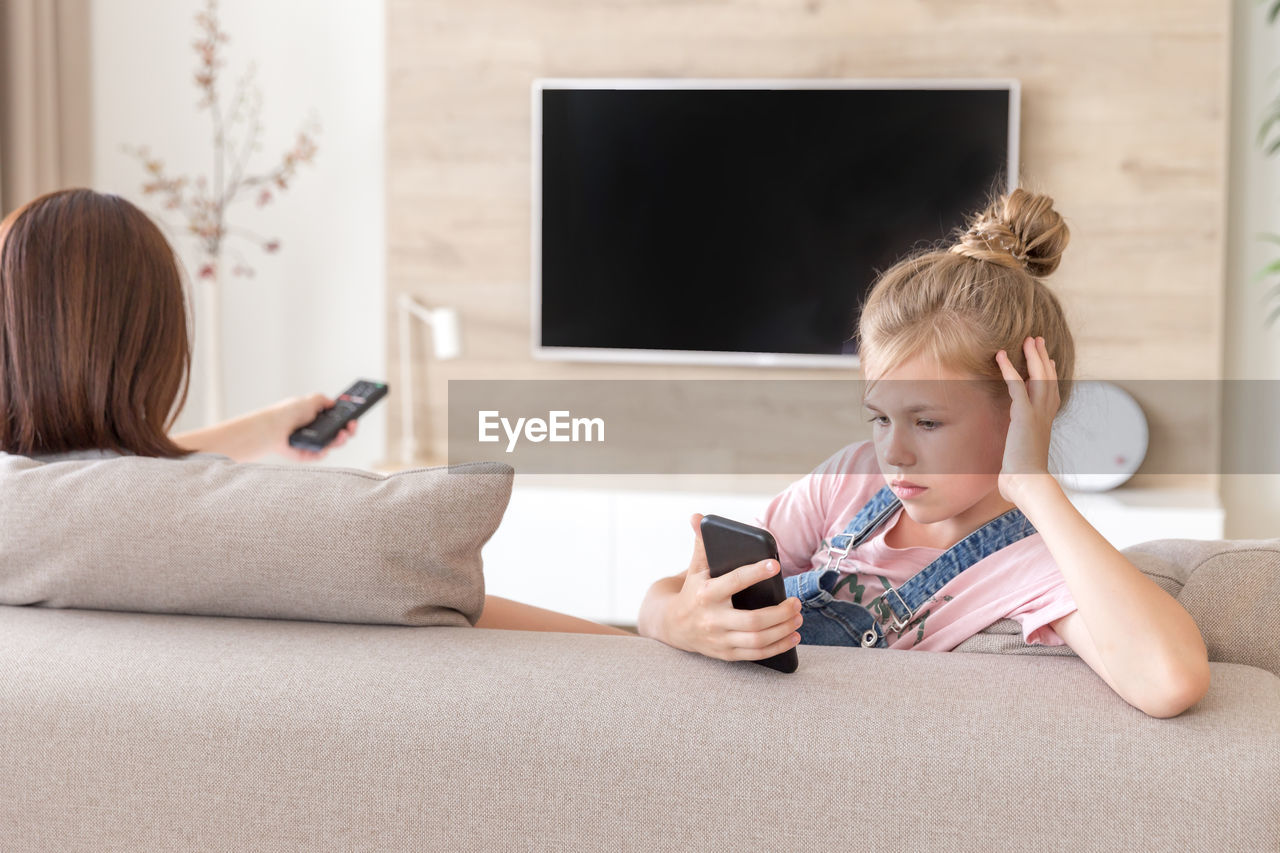 Girl looking at smart phone sitting by mother on sofa at home