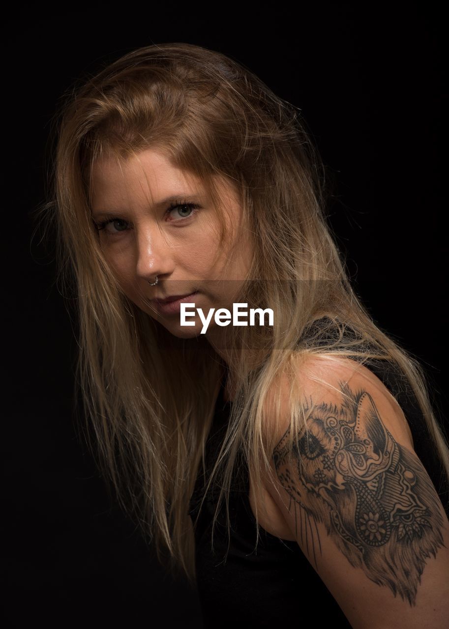 Close-up portrait of beautiful woman with tattoo on arm against black background