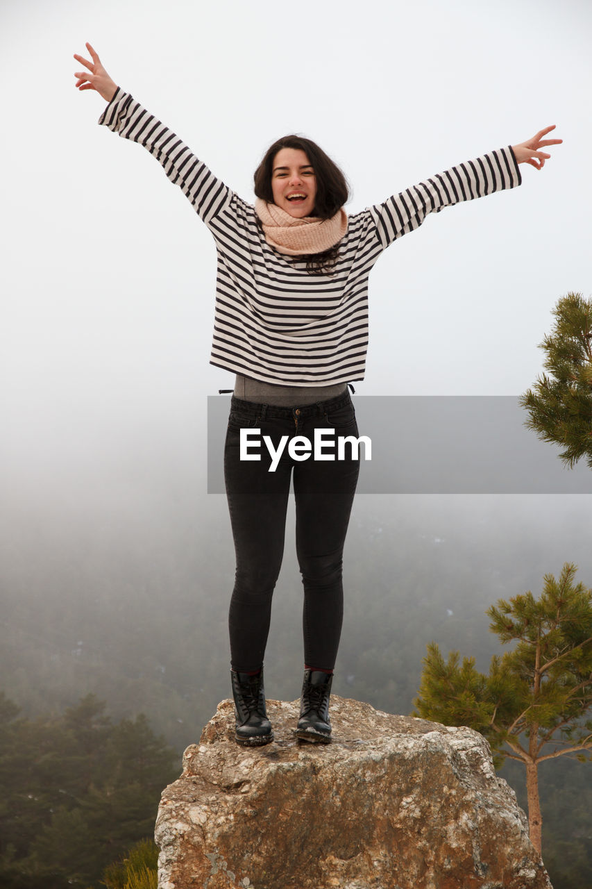 Full length of carefree young woman with arms raised standing on rock during foggy weather