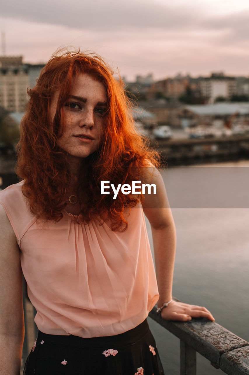 Portrait of young woman with red hair standing by railing on bridge