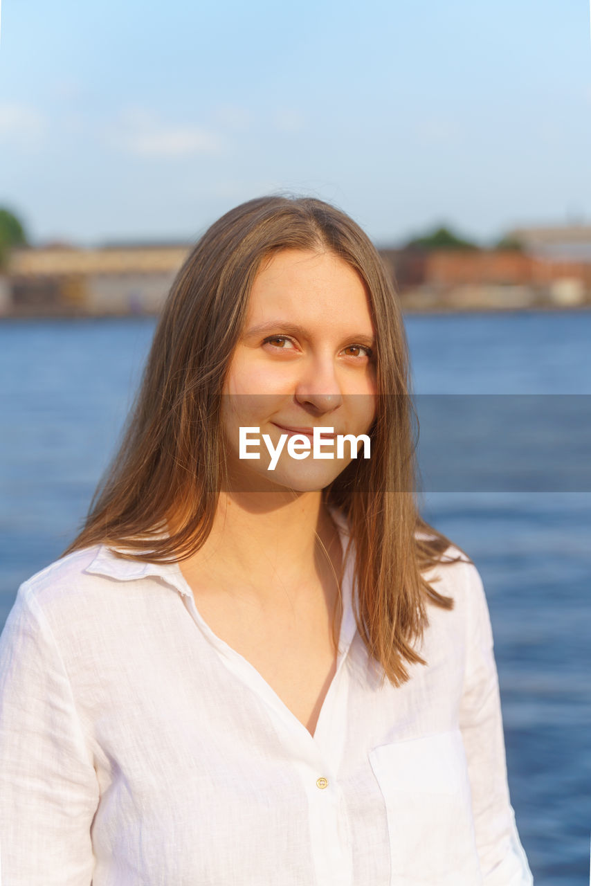 Portrait of cheerful young girl standing by river in city embankment, enjoing summer