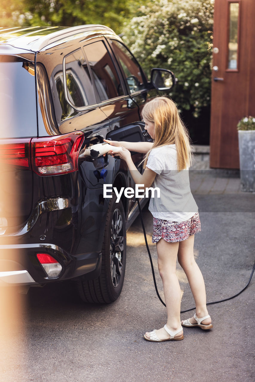 Blond girl charging black electric car in back yard