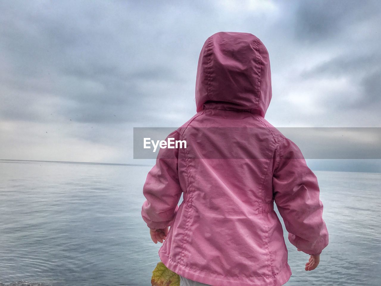 Rear view of baby girl in pink raincoat standing by lake geneva against cloudy sky