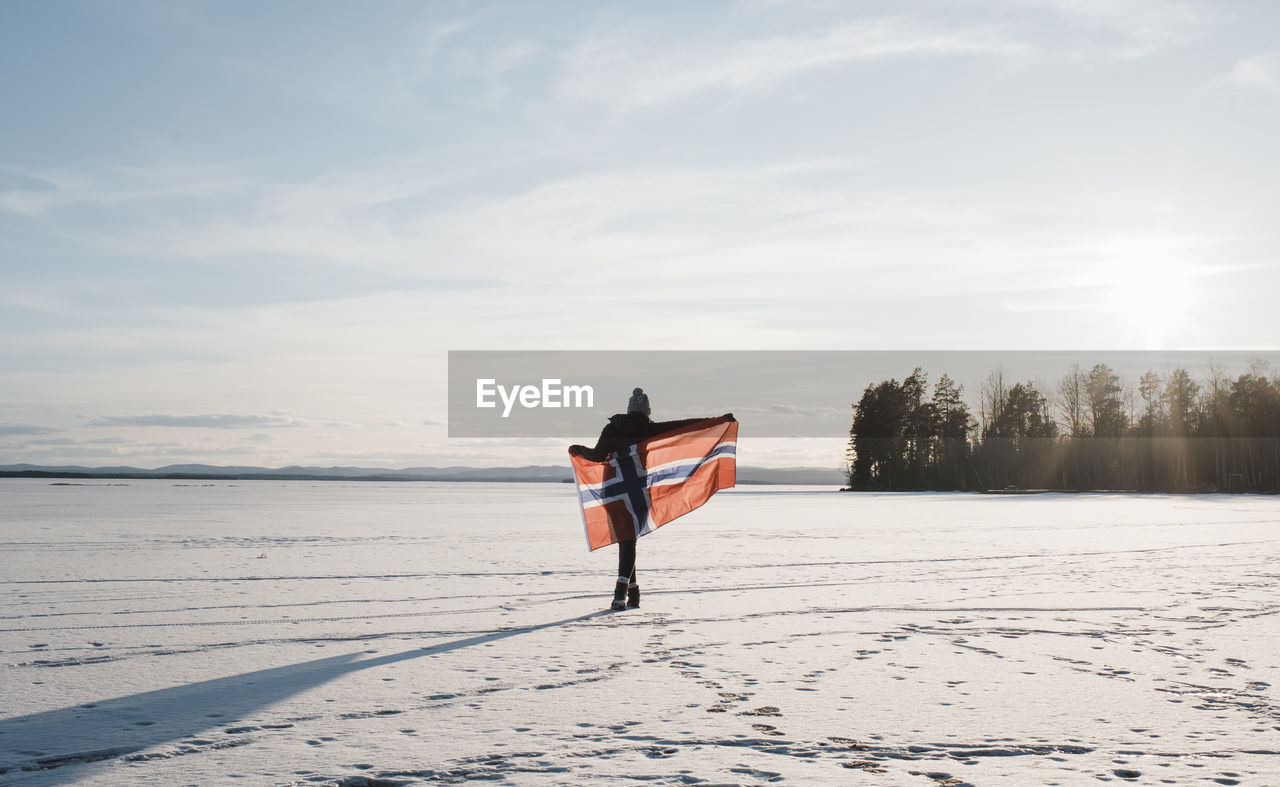 Woman walking across a frozen lake holding a norwegian flag at sunset