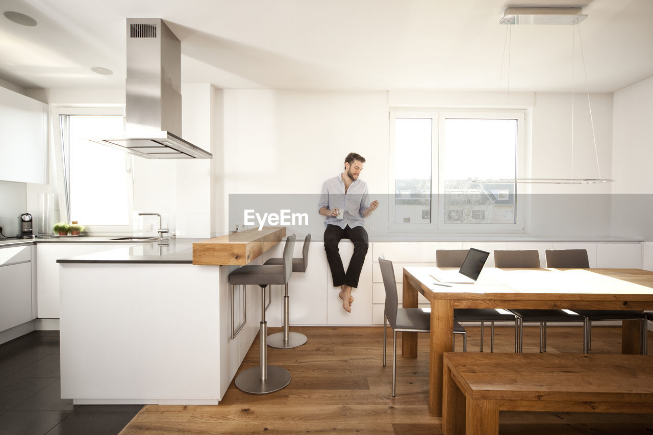 Man with coffee cup sitting on sideboard in his open plan kitchen looking at smartphone