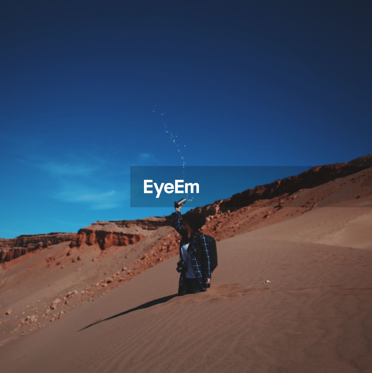 Woman throwing water while standing in sand against blue sky