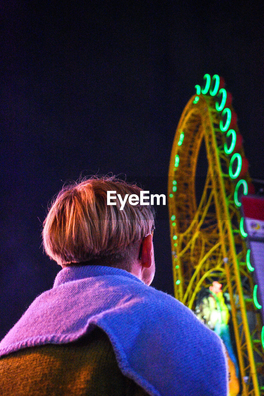 Low angle view of man standing against illuminated amusement park ride at night