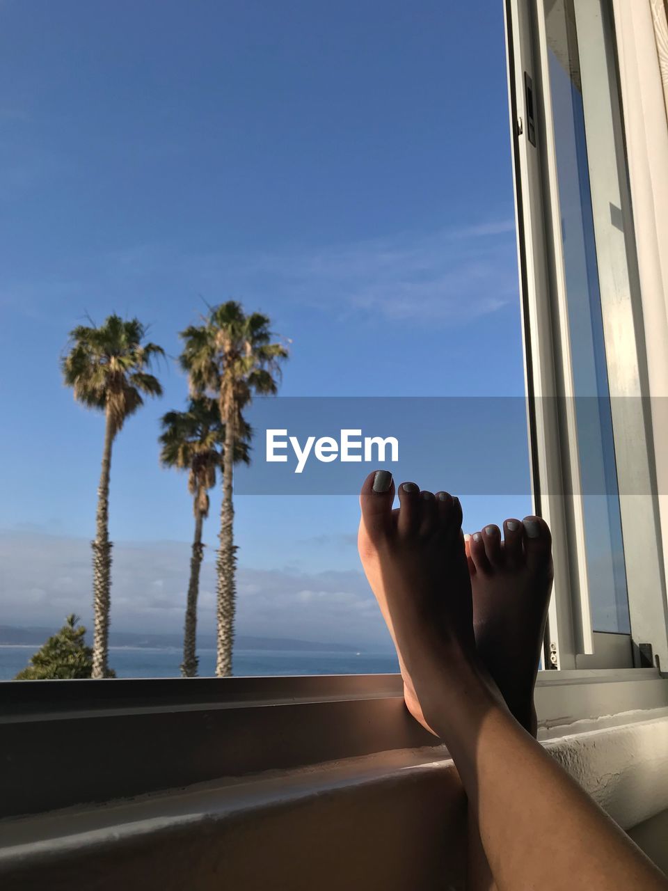 Low section of woman with feet on window sill against blue sky