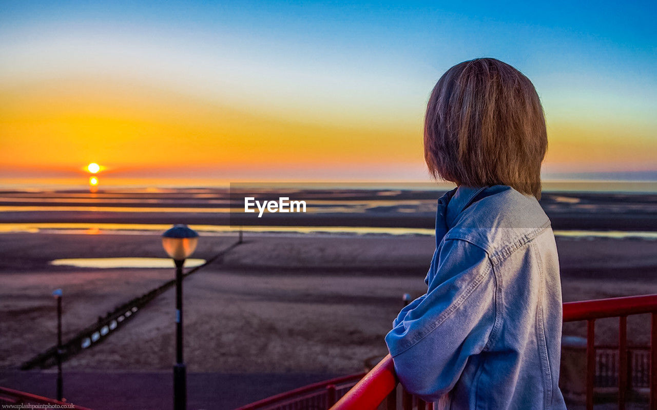 sky, sunset, one person, evening, blue, horizon, sea, nature, water, sunlight, rear view, women, beauty in nature, transportation, dusk, railing, waist up, ocean, adult, childhood, beach, standing, child, reflection, outdoors, cloud, looking at view, tranquility, looking, lifestyles, casual clothing, land, scenics - nature, travel, vacation, person, travel destinations, leisure activity, orange color, tranquil scene, trip, focus on foreground, hairstyle