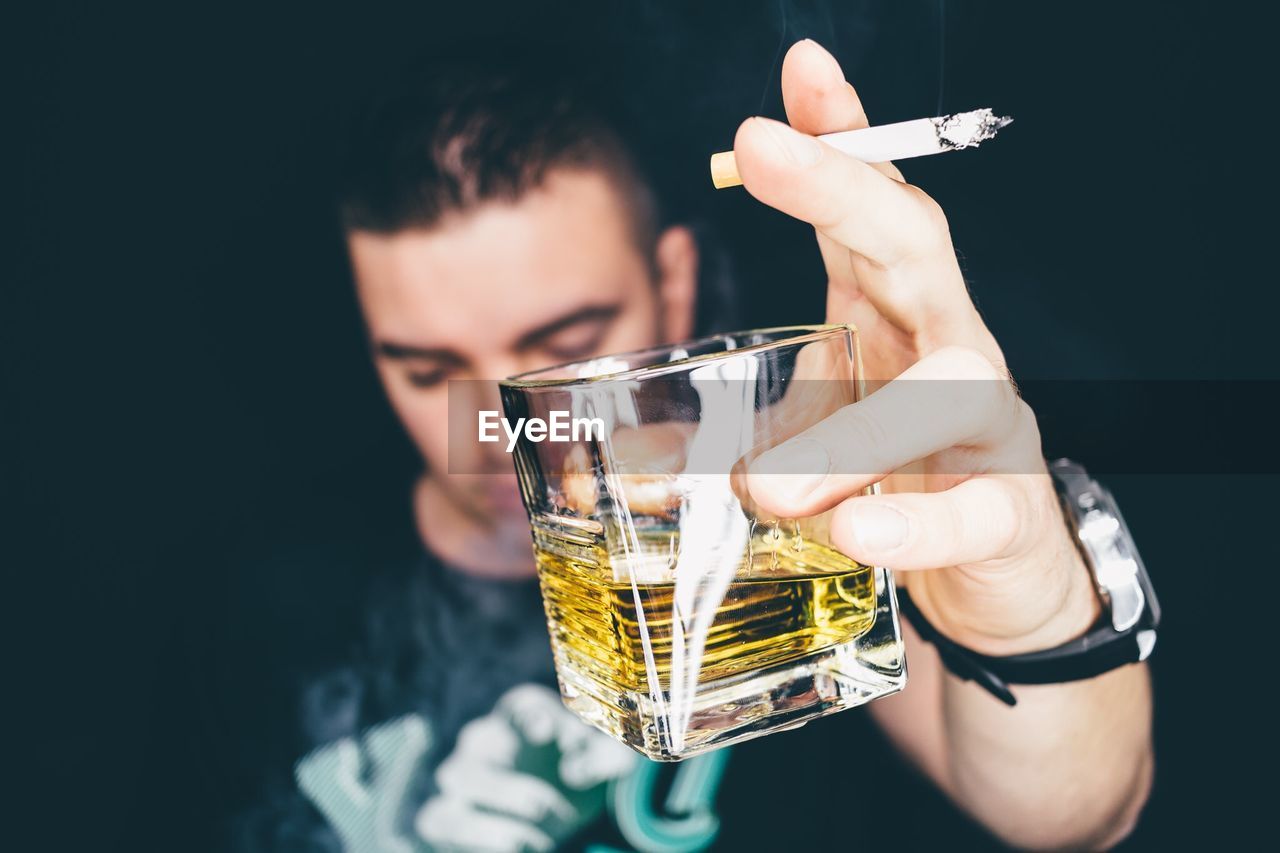 Close-up of man holding cigarette and whiskey glass against black background