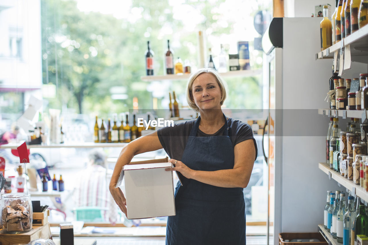 Portrait of confident female owner carrying cardboard box in deli
