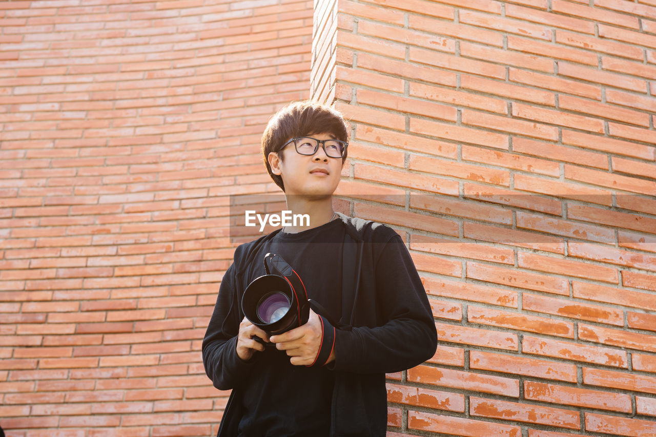 A young asian man stands in front of a brick wall holding a camera and looking forward.