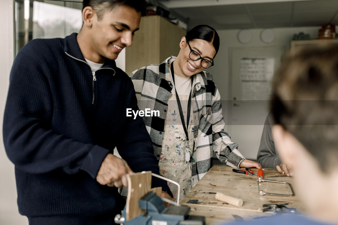 Happy female teacher standing with male student cutting wood during carpentry class