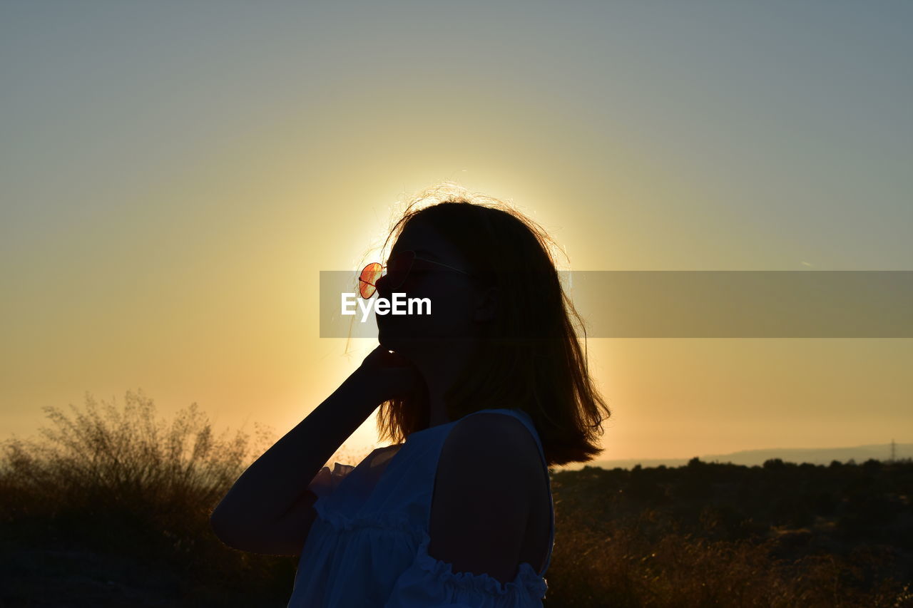 WOMAN STANDING ON FIELD AGAINST SKY DURING SUNSET