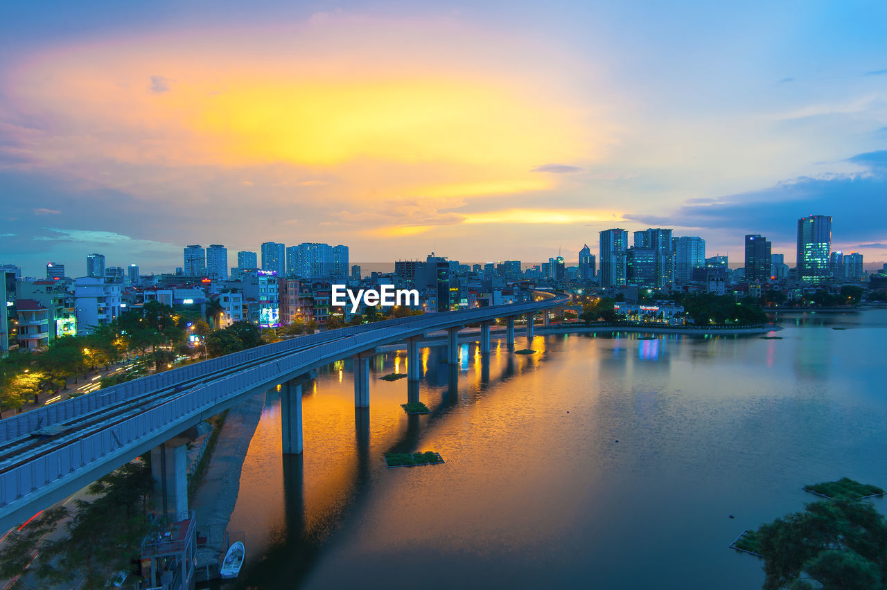 Illuminated buildings by river against sky during sunset