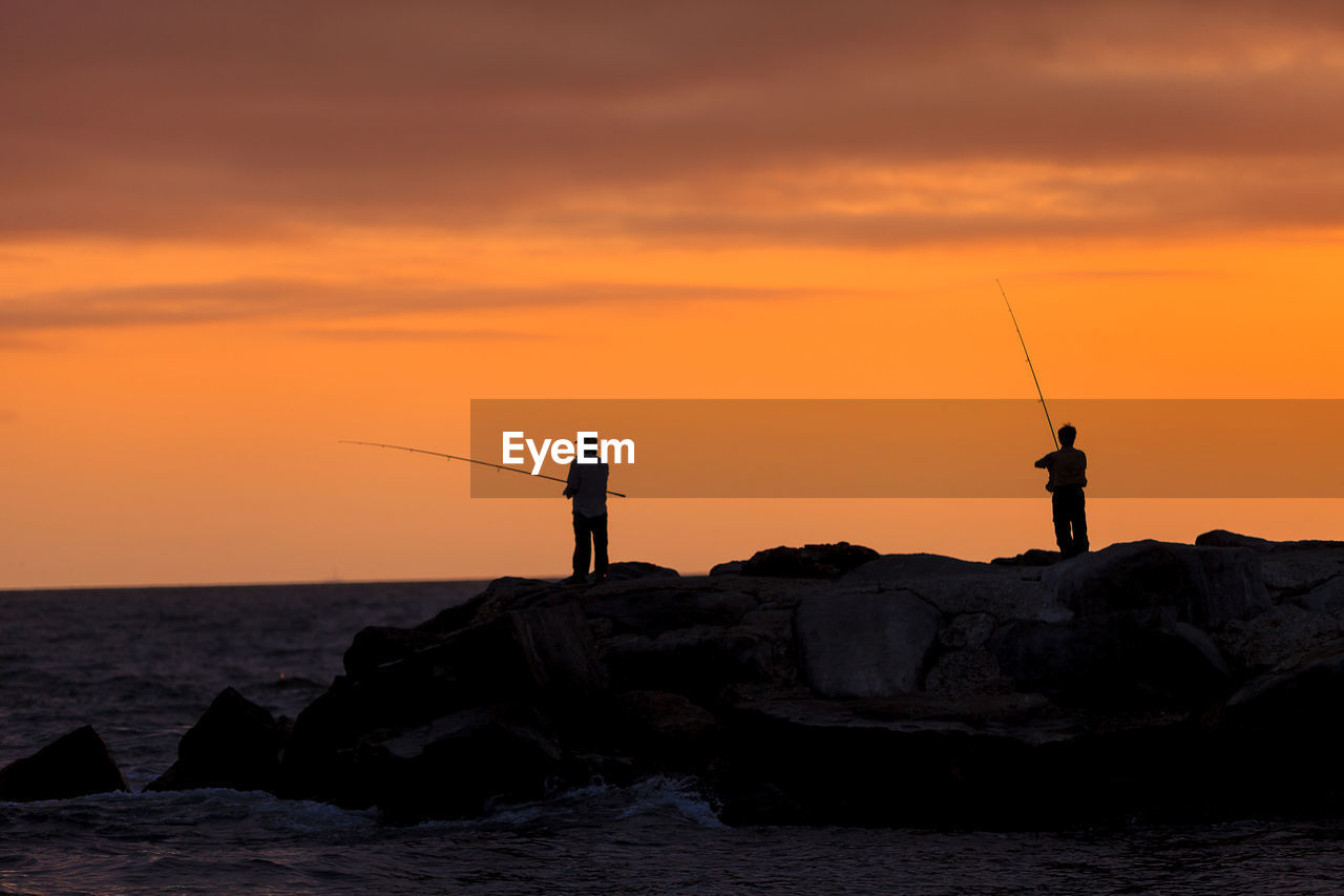 SILHOUETTE MAN FISHING ON ROCK AGAINST SEA DURING SUNSET
