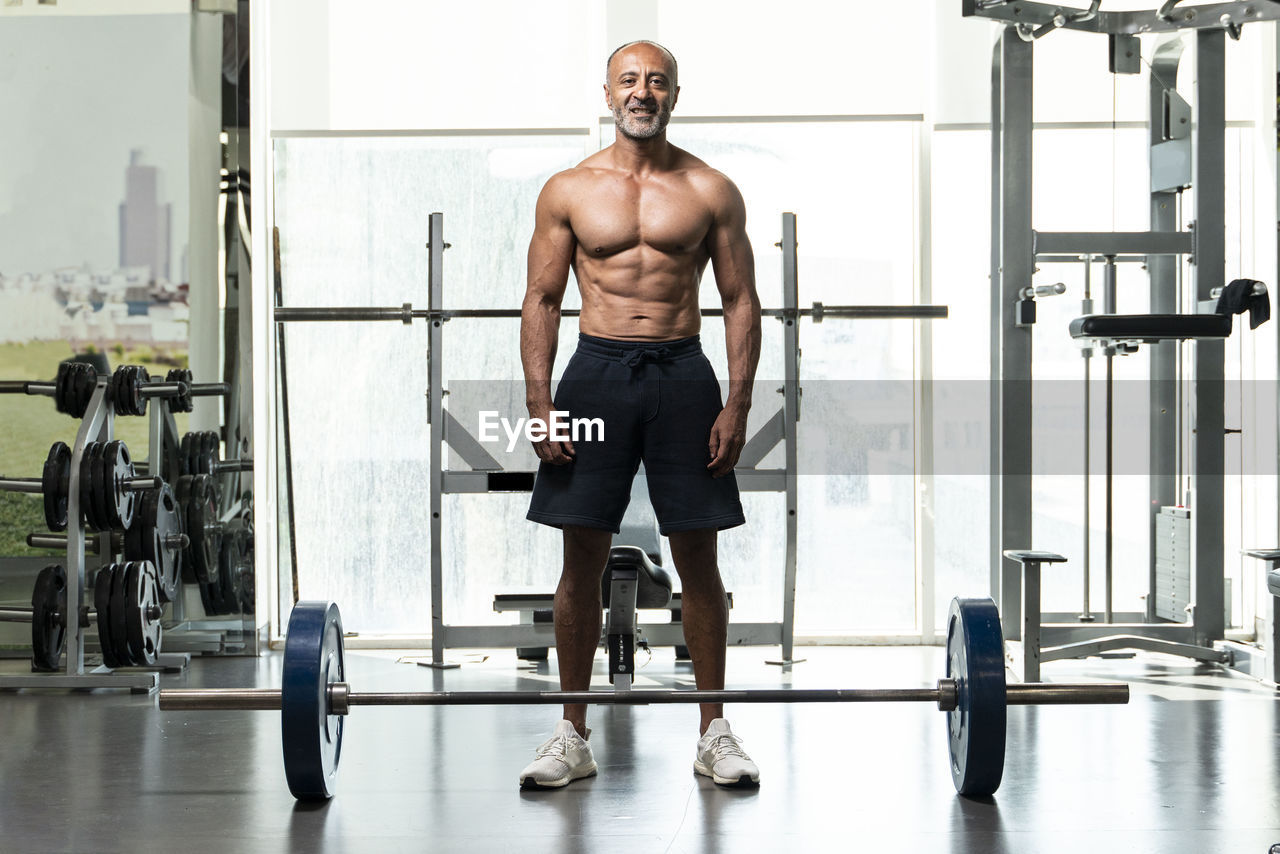 Full length of shirtless man with barbell standing in gym