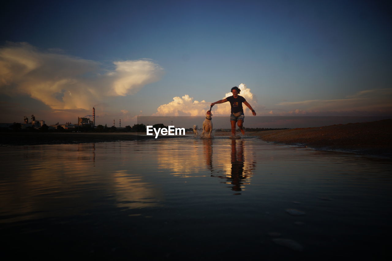 Woman with dogs running on shore at beach against sky during sunset