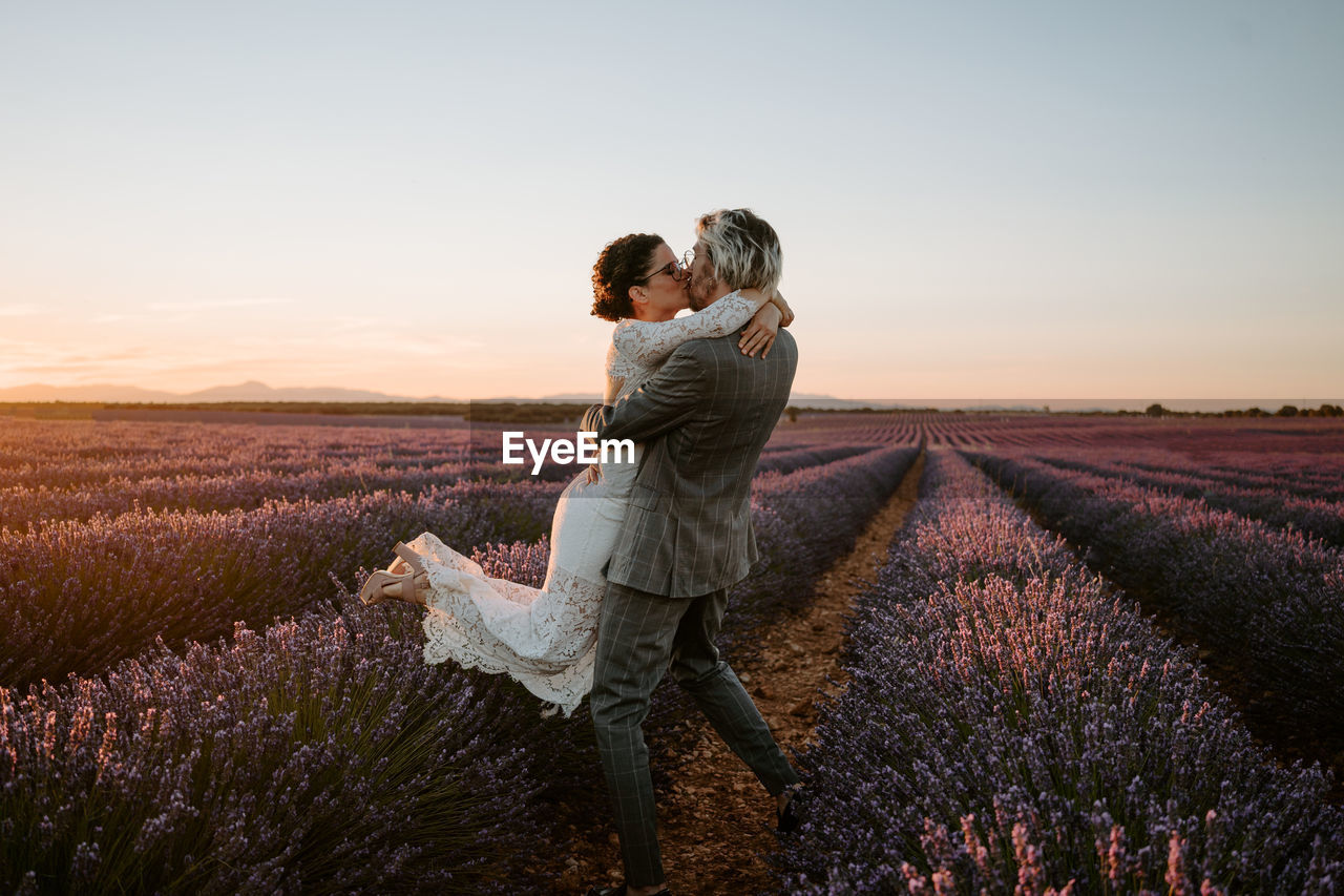 Side view of groom lifting bride while standing in lavender field on background of sunset sky on wedding day