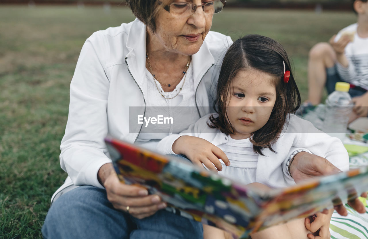 Grandmother showing picture book to granddaughter on meadow