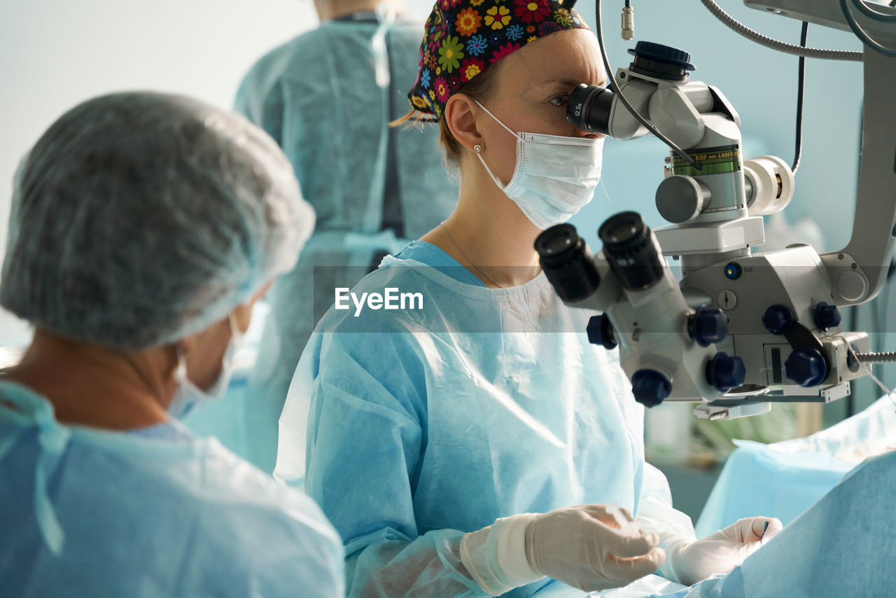 Adult female doctor in sterile mask and ornamental medical cap looking through surgical microscope against crop coworker in hospital