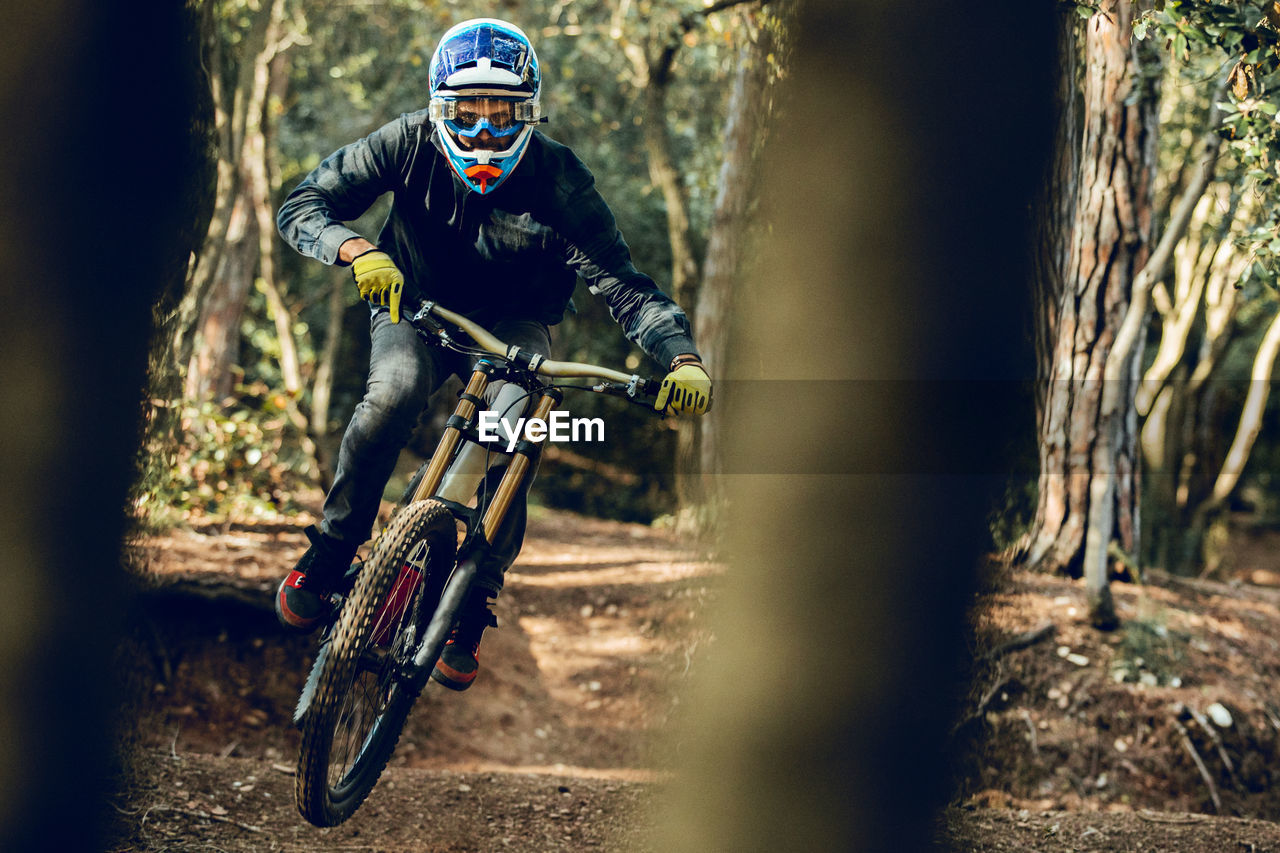 Unrecognizable man looking at camera in helmet, gloves and protection glasses jumping doing whip trick downhill during mountain biking practice in wood forest