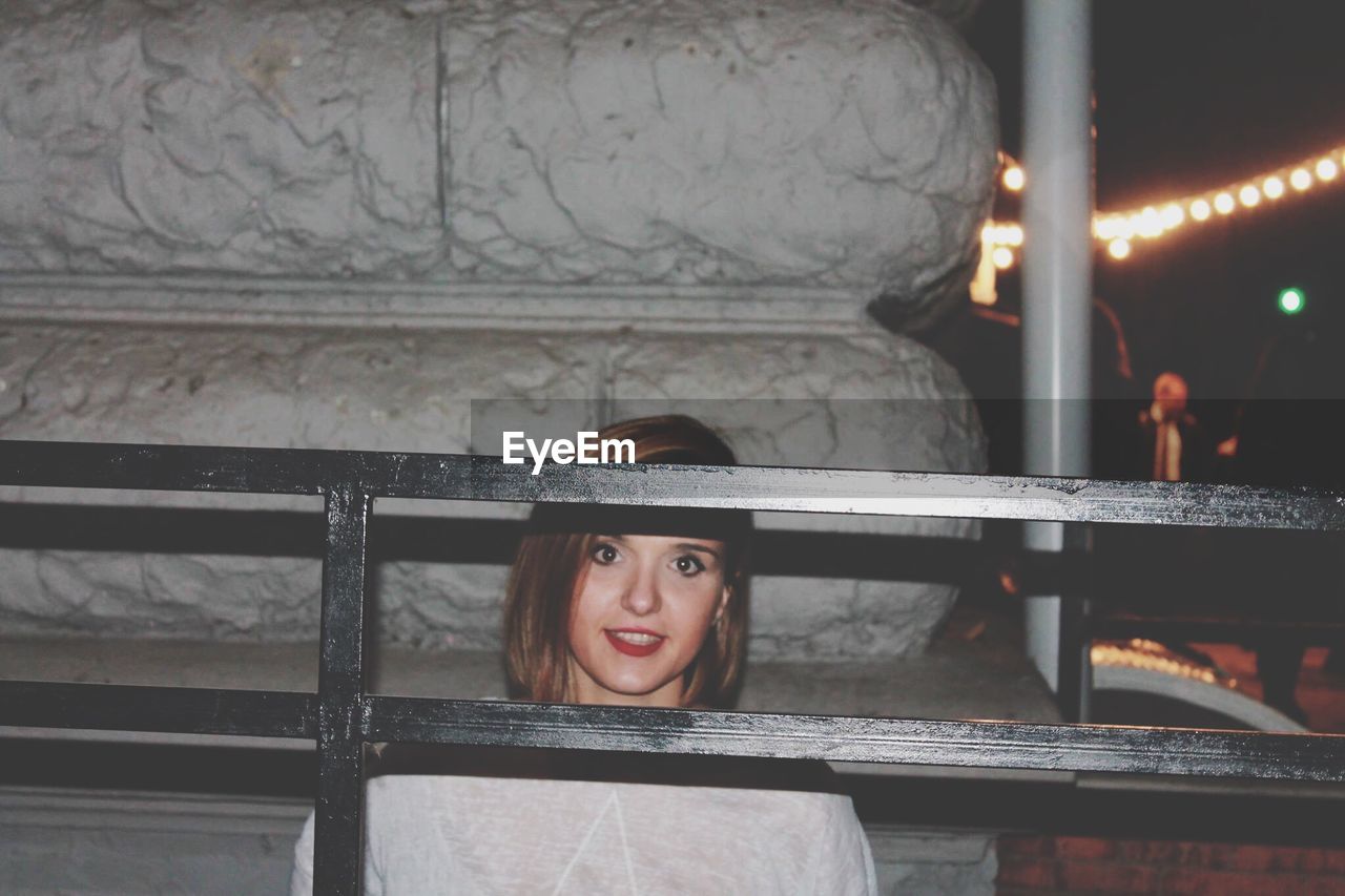 Close-up portrait of young woman seen through railing at night