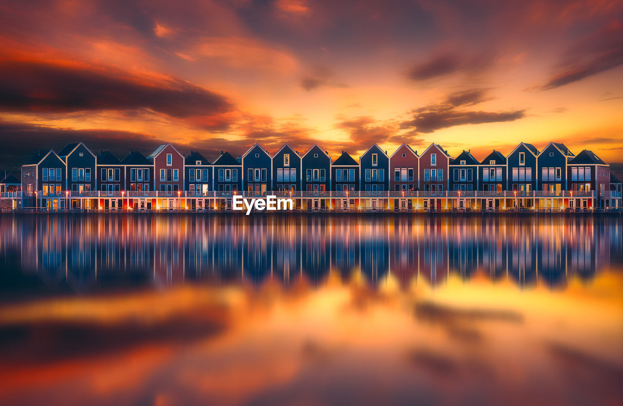 Houses reflecting on calm lake against orange sky during sunset