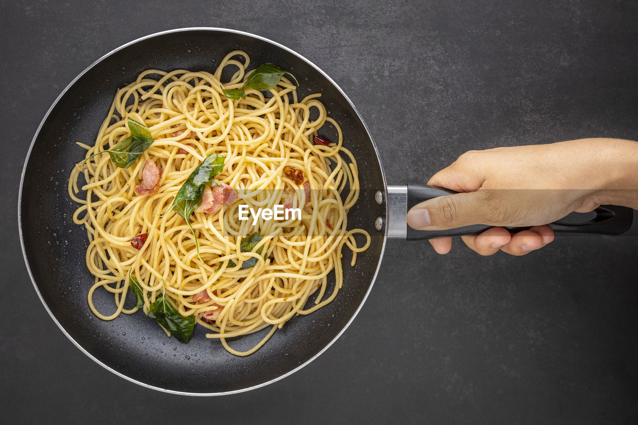 Hand holding pan of spaghetti pasta with dried chilli, garlic, sweet basil and bacon, top view